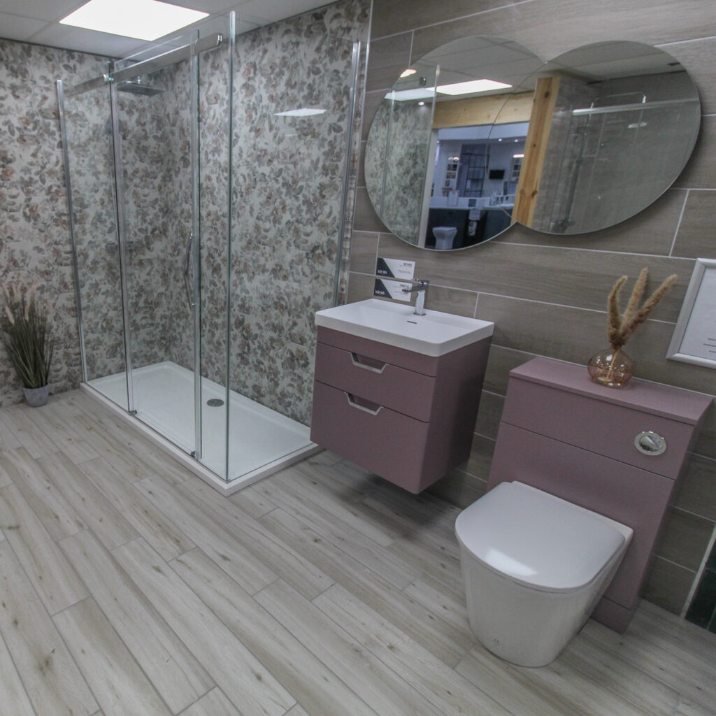 Contemporary bathroom display with walk-in glass shower, pink vanity unit, matching WC unit, and floral feature wall at Coventry Plumbing and Heating Supplies showroom.